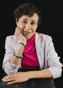 promo portrait of Kelly Bron Johnson, a light-skinned Black person with short curly dark brown hair brushed to one side. Kelly is wearing a light pink blazer over a fuchsia turtleneck shirt. Her face is leaning on her right hand and she is looking directly at the camera with a small, closed lipped smile.