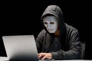 Person dressed in dark hoodie with hood pulled up wearing a white mask while typing on a laptop keyboard