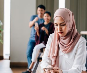 Woman wearing a hijab looking at her phone with a concerned expression on her face while two people in the background point at her and laugh