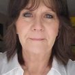 An older white woman with brown hair wearing a white shirt smiles into the camera.